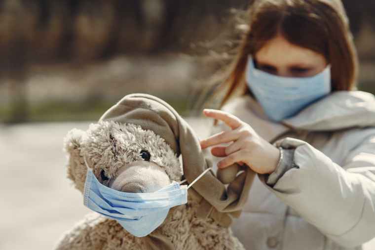 cute girl in medical mask wearing mask on teddy bear during pandemic